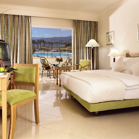 Movenpick Taba Resort & Spa Oda fotoğraf The photo shows an elegant hotel room featuring a large bed with white linens and several pillows. There is a small dining table with green chairs beside it and a television on a wooden unit. Large windows with sheer curtains let in natural light, of