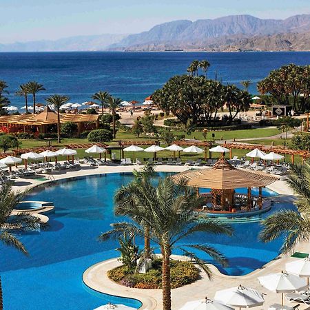 Movenpick Taba Resort & Spa Dış mekan fotoğraf The photo shows a luxurious resort overlooking a large, blue swimming pool. Surrounding the pool are palm trees and lounge chairs with white umbrellas, creating a relaxing atmosphere. In the background, the ocean can be seen, with a mountainous lands
