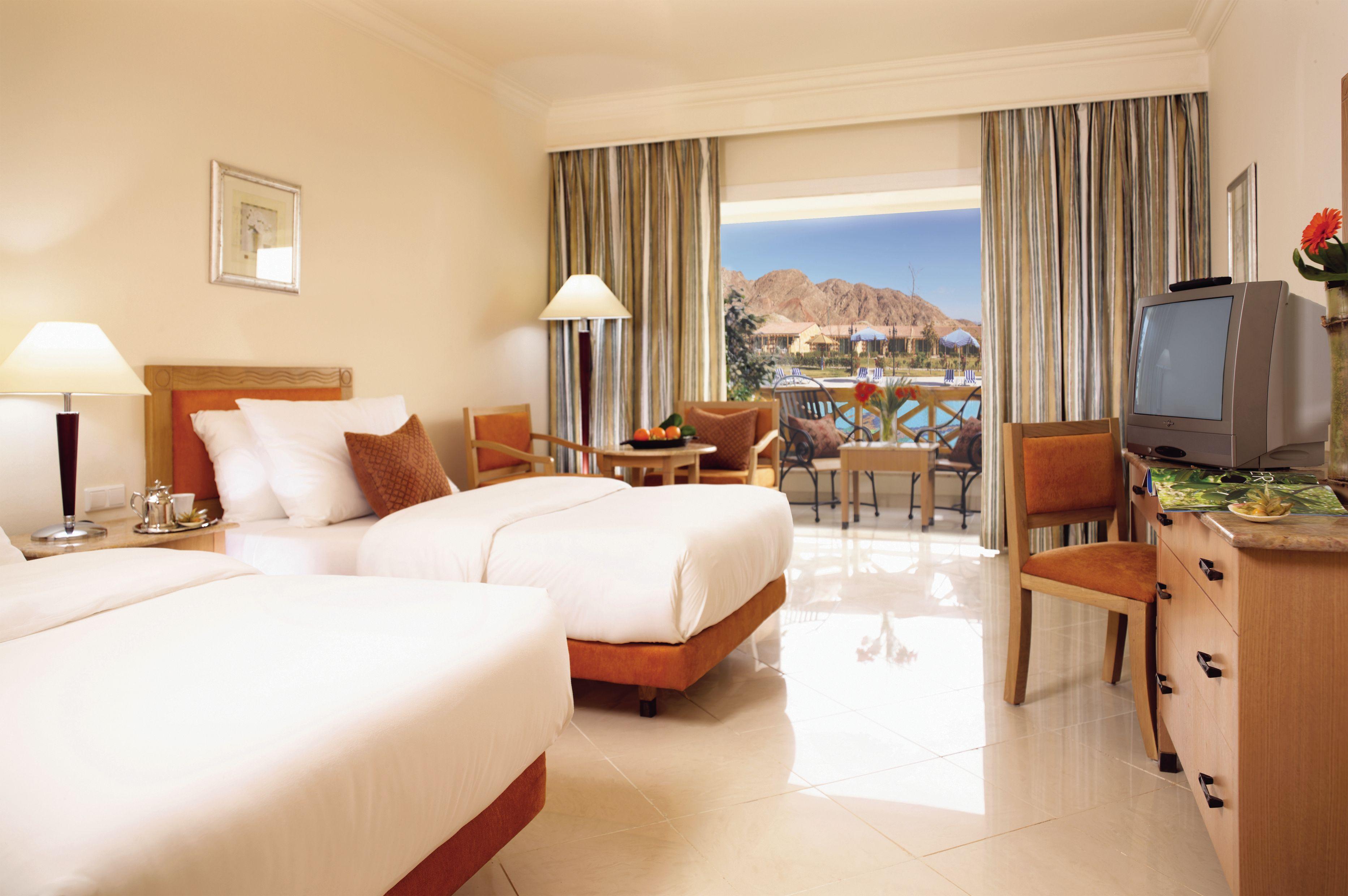 Movenpick Taba Resort & Spa Oda fotoğraf The photo shows a hotel room with two twin beds placed side by side. The bedding is light in color, and there are decorative pillows on each bed. A wooden bedside table with a lamp is positioned next to one of the beds. 

In the background, there are