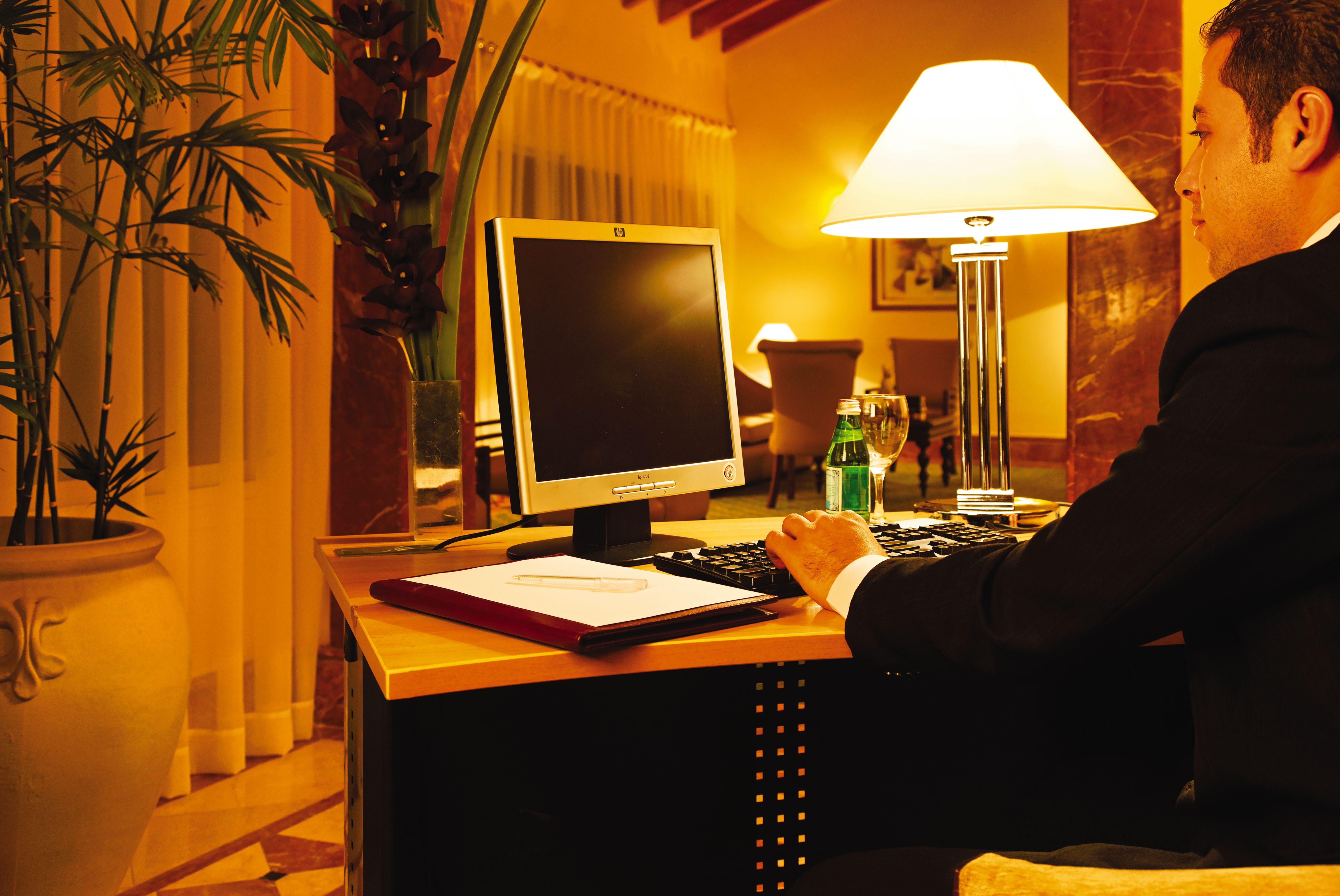 Movenpick Taba Resort & Spa Dış mekan fotoğraf The photo shows a workspace with a desk and a computer. The desk is positioned in a well-lit area, with soft lighting coming from a nearby lamp. A person, dressed in a suit, is seated at the desk, using the computer. Next to the computer, there is a 