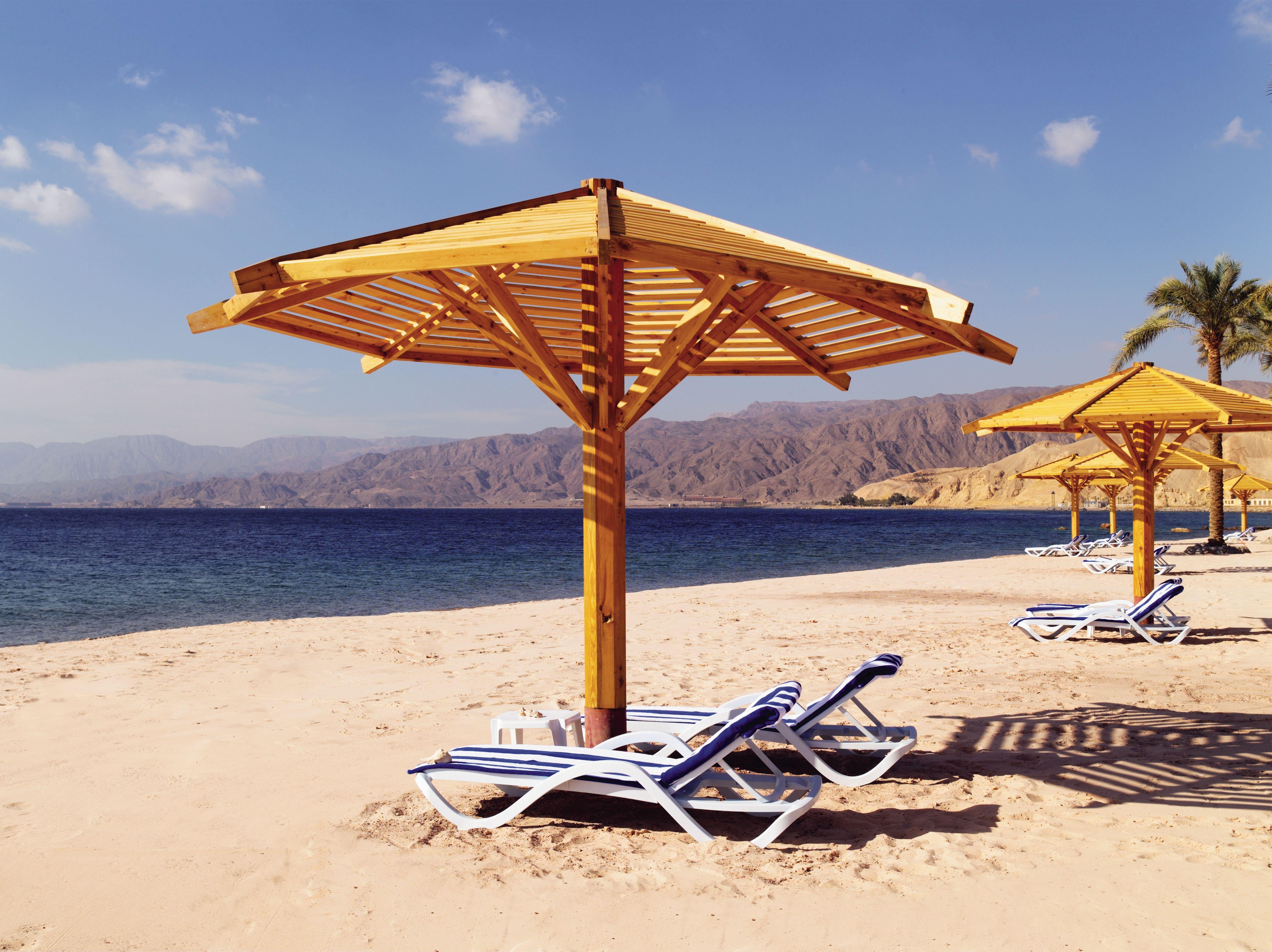 Movenpick Taba Resort & Spa Dış mekan fotoğraf The photo depicts a tranquil beach scene. There are several wooden umbrellas providing shade over white lounge chairs, arranged on the sandy beach. In the background, you can see a calm sea with gentle waves and distant mountains under a clear blue s