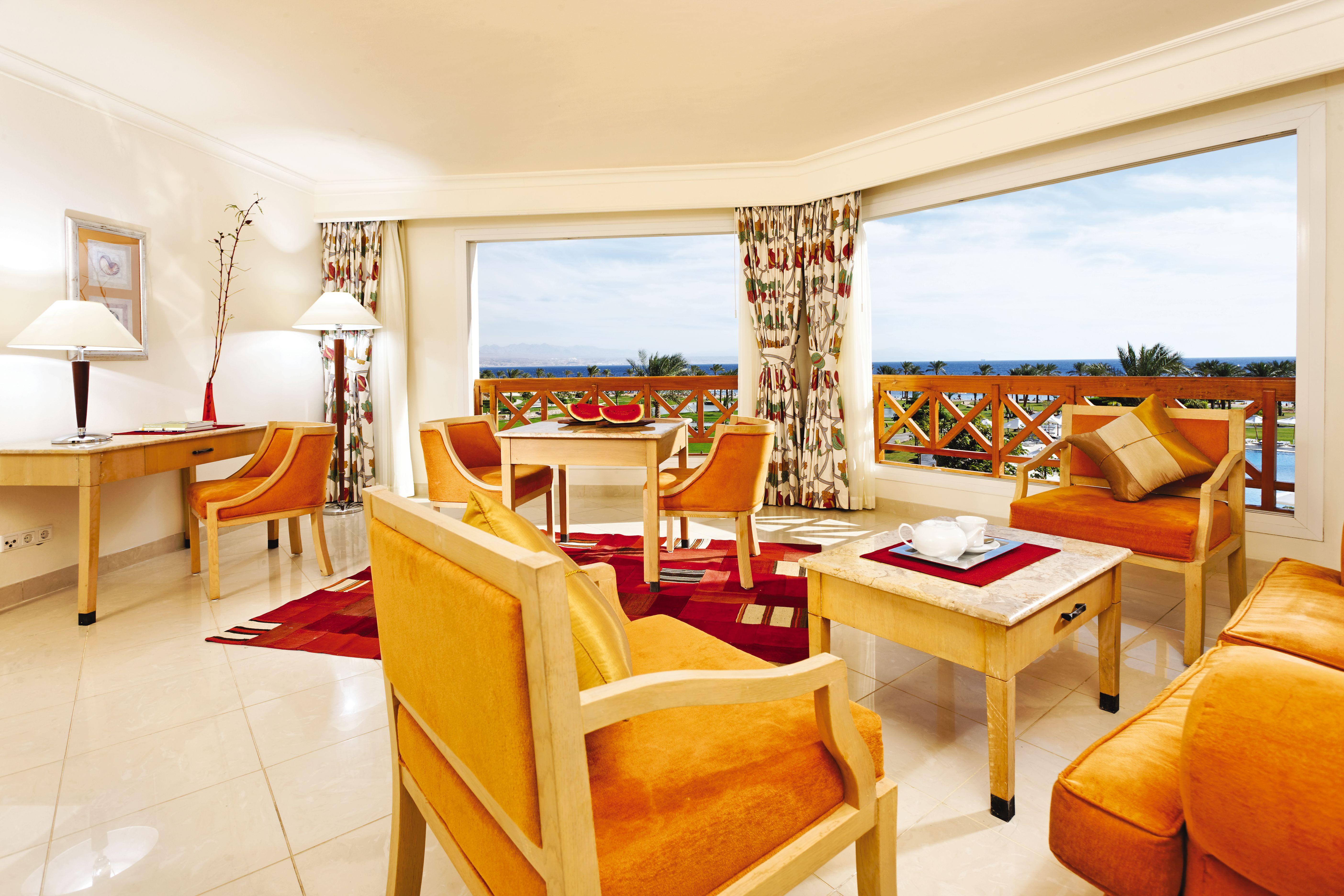 Movenpick Taba Resort & Spa Dış mekan fotoğraf The photo shows a bright and upscale hotel suite. It features a spacious living area with large windows that offer a scenic view outside. The decor includes warm-toned furniture, such as orange armchairs and wooden tables. There is a small dining tab