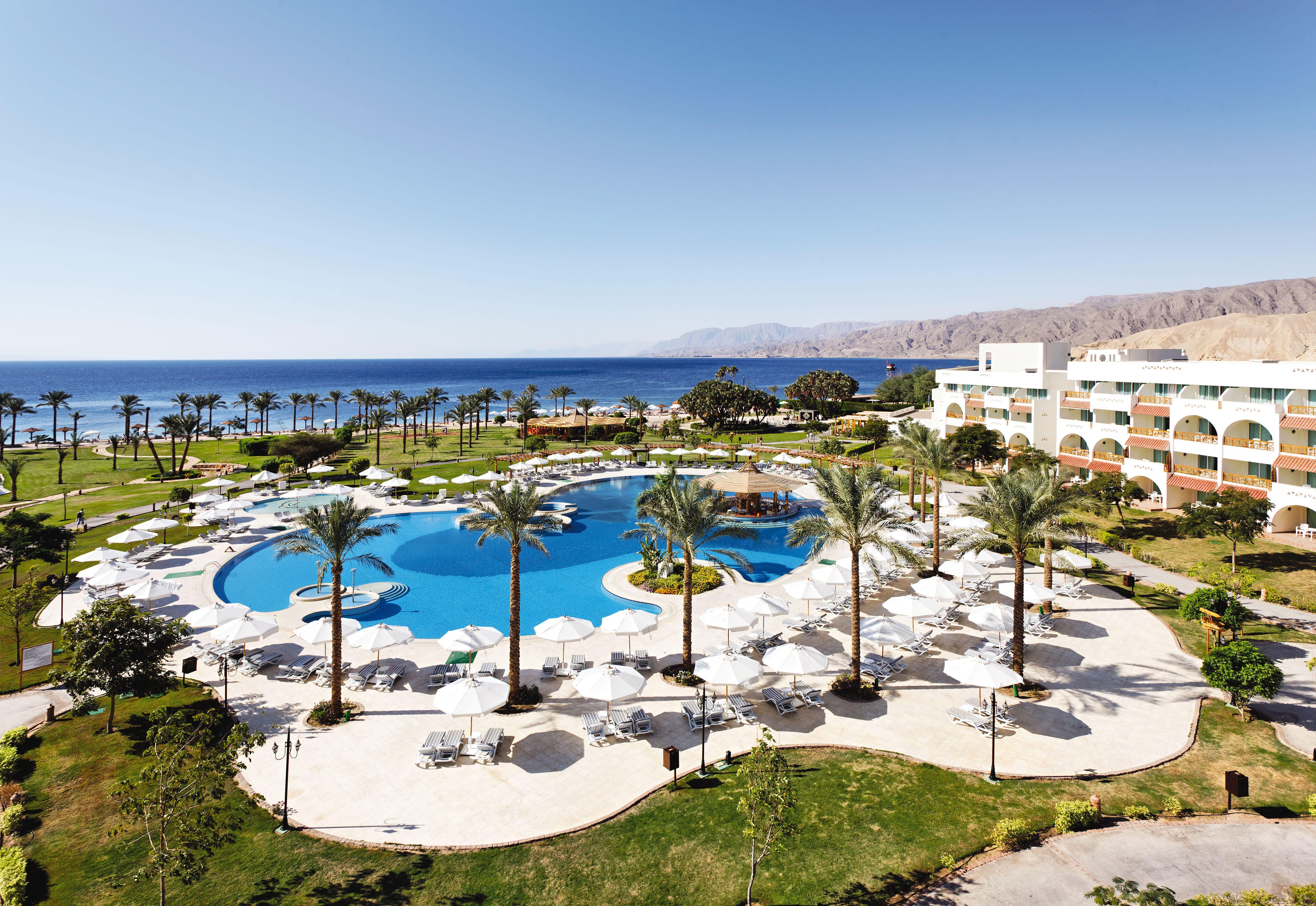 Movenpick Taba Resort & Spa Oda fotoğraf The photo shows a scenic view of a resort property. In the foreground, there is a large swimming pool surrounded by lush greenery and palm trees. Lounge chairs and umbrellas are neatly arranged around the pool area, suggesting a relaxing atmosphere. 