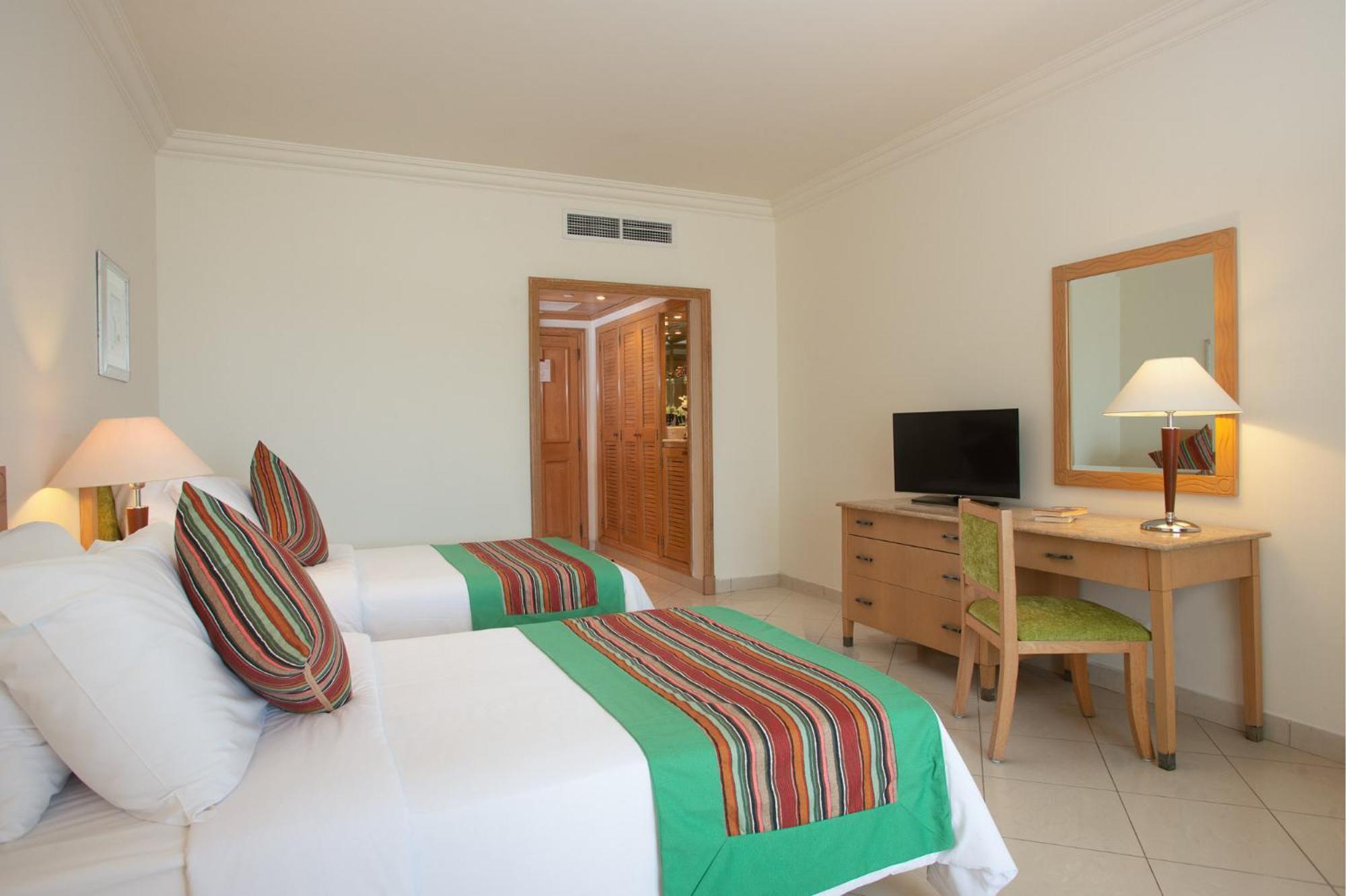 Movenpick Taba Resort & Spa Dış mekan fotoğraf The photo shows a hotel room featuring two beds with colorful striped throw blankets. There are decorative pillows on each bed, and the overall decor appears bright and modern. A wooden desk with a chair is positioned next to a TV, and there is a mir