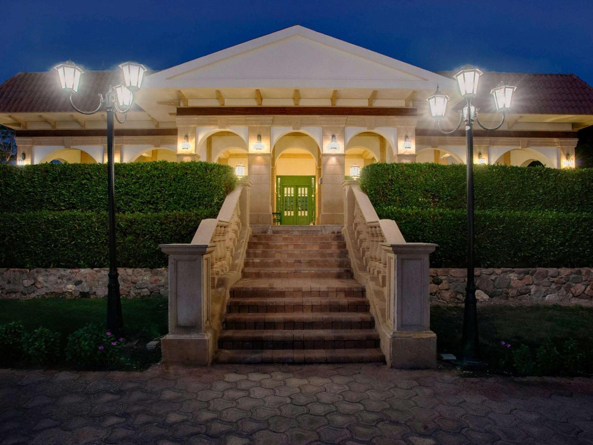 Movenpick Taba Resort & Spa Dış mekan fotoğraf The photo shows a large house or villa with a grand entrance. The structure features a sloped roof and is adorned with columns and detailed architectural elements. There are steps leading up to a double-door entry that is painted green. 

On either s