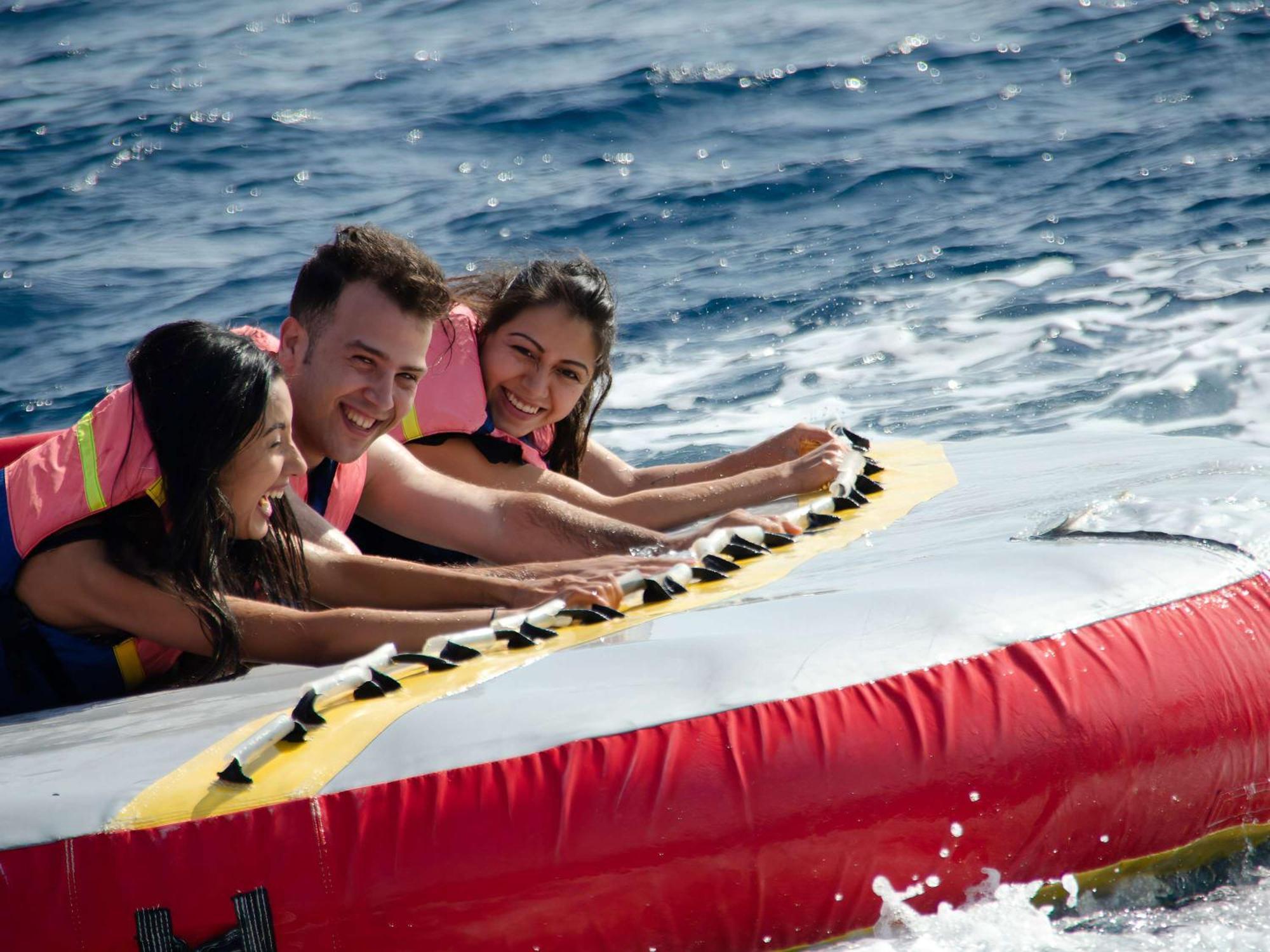 Movenpick Taba Resort & Spa Dış mekan fotoğraf The photo shows three people enjoying a fun activity on a water inflatable. They are riding a banana boat, which is being pulled by a speedboat on the water. The individuals are wearing life jackets, and they all appear to be smiling and having a gre