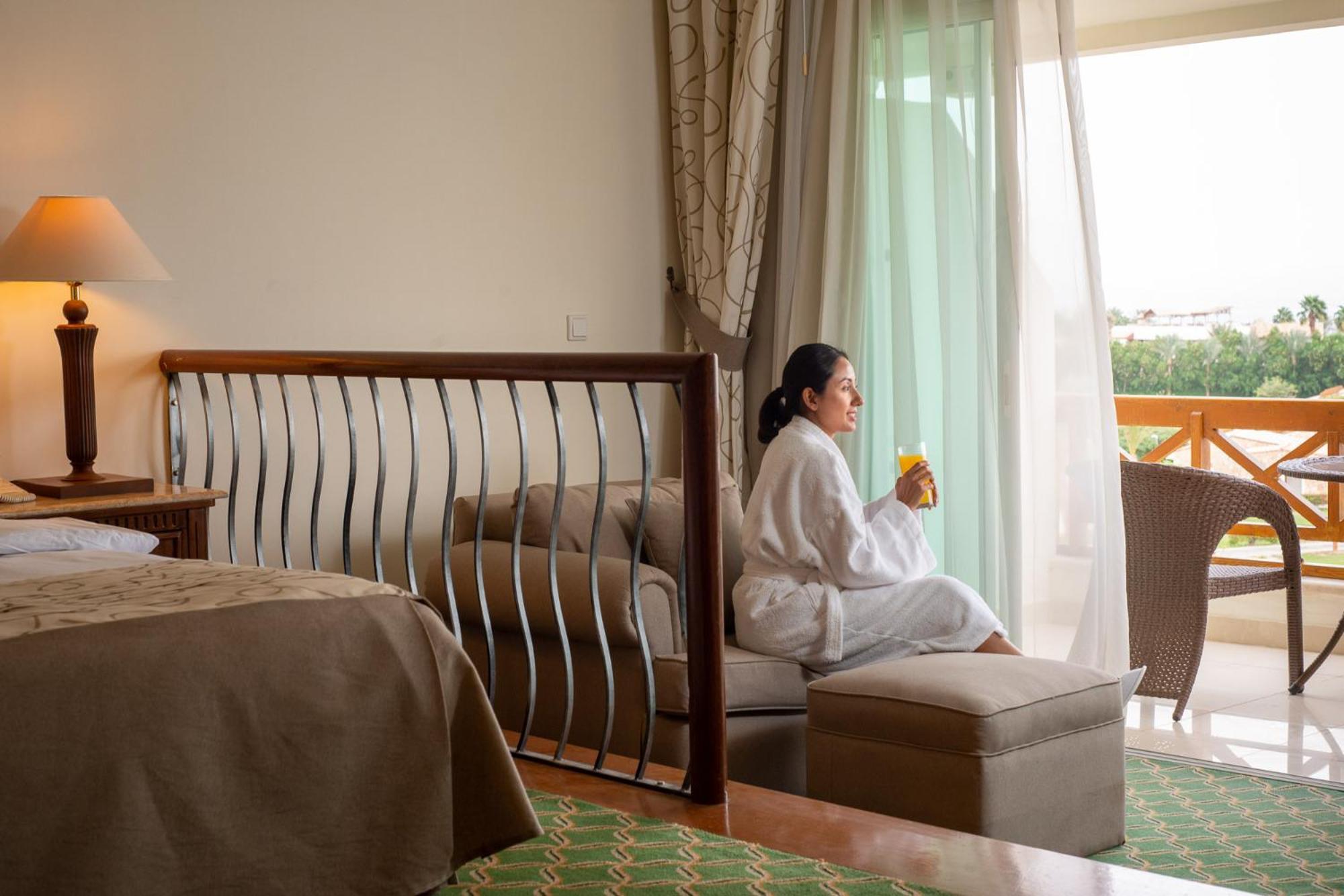 Movenpick Taba Resort & Spa Dış mekan fotoğraf The photo shows a cozy hotel room with elegant decor. In the foreground, there is a bed with a neutral-colored blanket and a stylish bed frame with decorative metalwork. A woman in a white bathrobe is sitting on a small ottoman, enjoying a drink. She