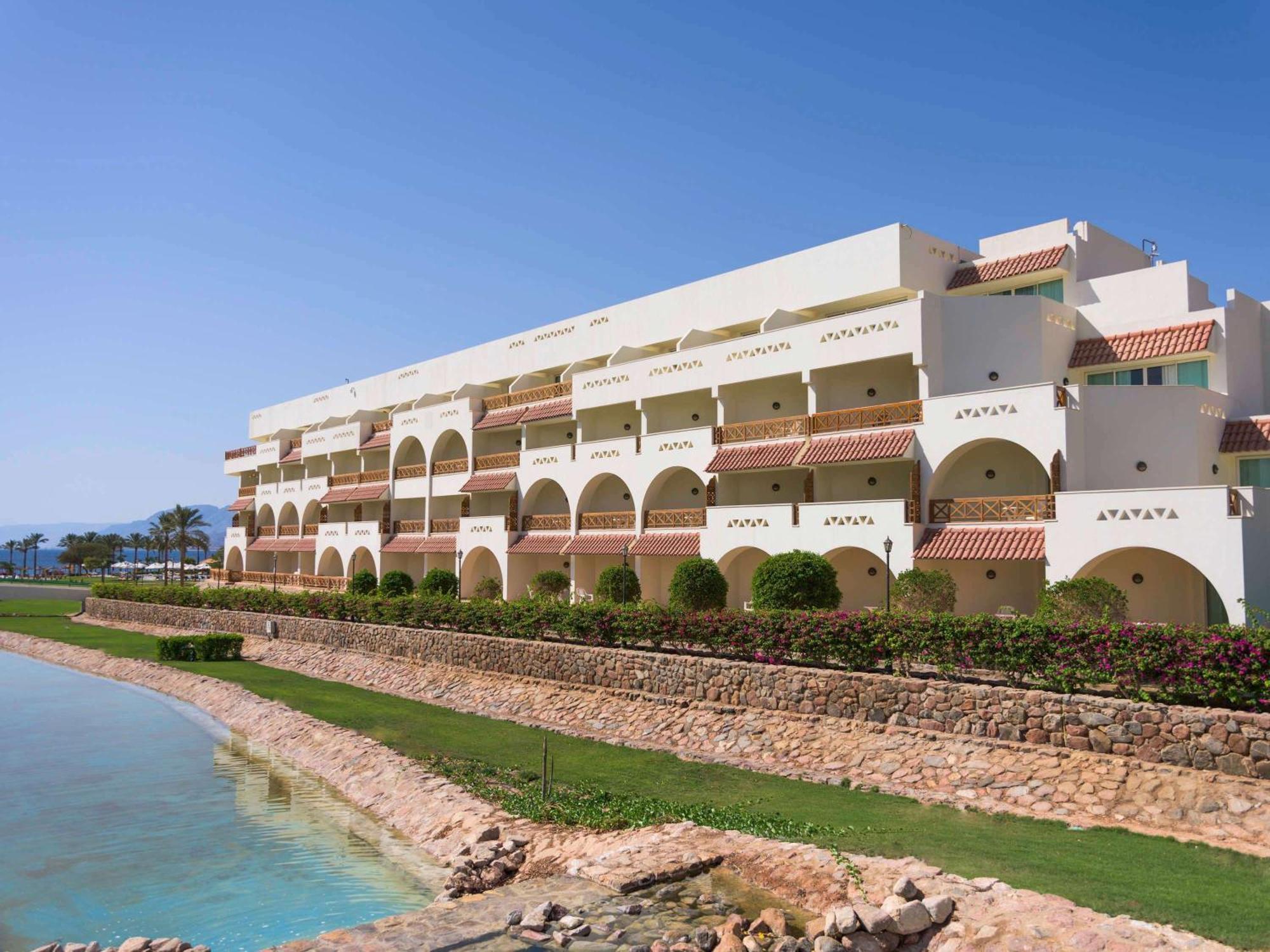 Movenpick Taba Resort & Spa Dış mekan fotoğraf The photo shows a modern, multi-story building with a white and light-colored exterior design. It features multiple arches and balconies, giving it a Mediterranean architectural style. The building is situated near a body of water, possibly a lagoon 
