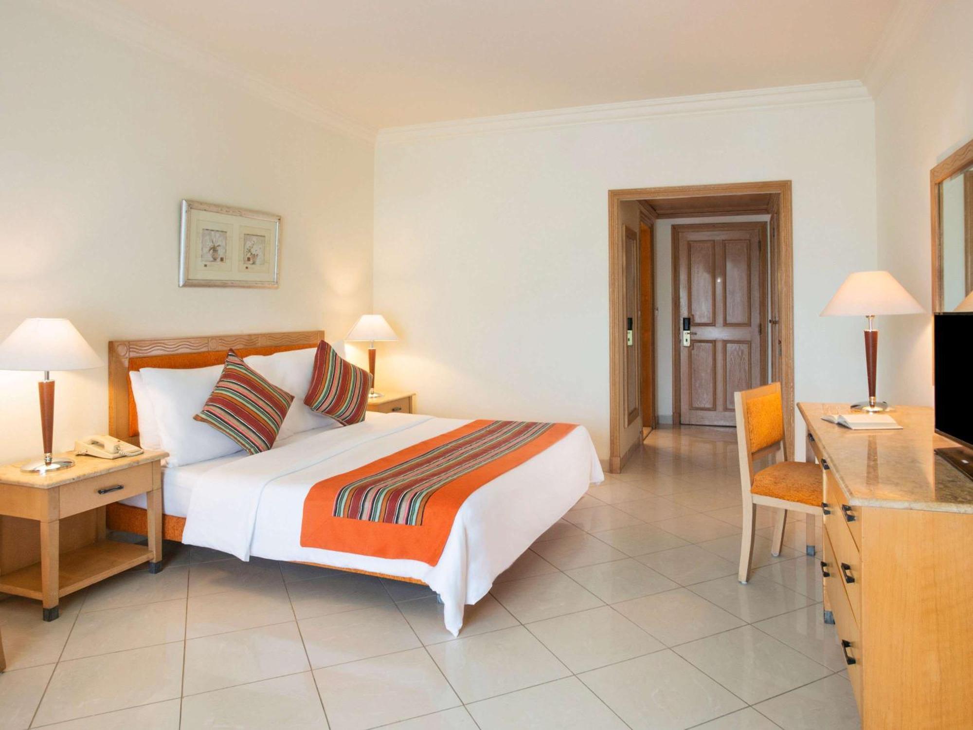 Movenpick Taba Resort & Spa Dış mekan fotoğraf The photo shows a well-lit hotel room that features a double bed with a white bedspread and decorative pillows in vibrant colors. There's an orange bed runner across the bed. The room has a simple and modern design, with tiled flooring. 

On one side