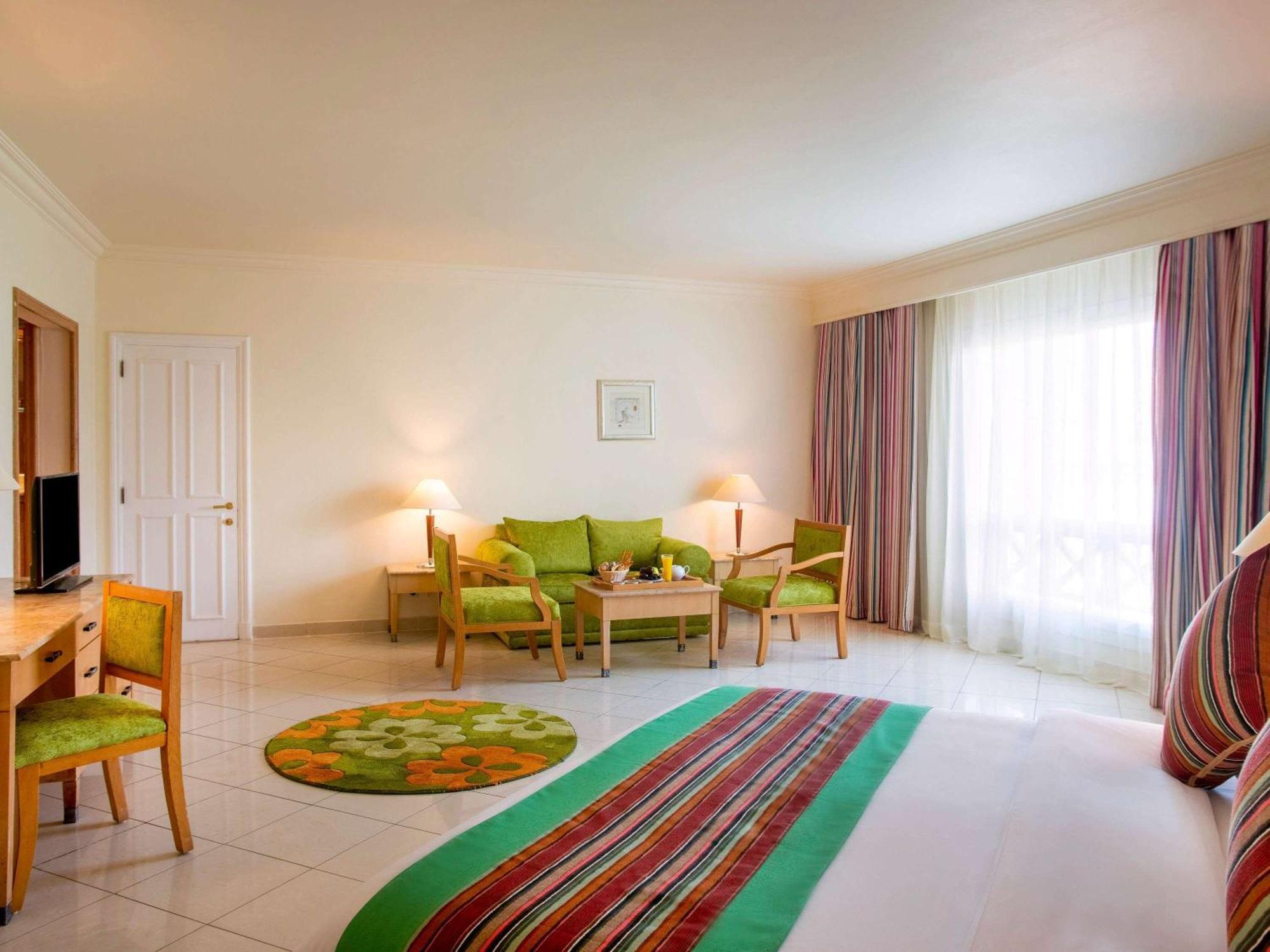Movenpick Taba Resort & Spa Dış mekan fotoğraf The photo shows a bright and airy hotel room or living area. It features a light-colored floor and walls, with large windows that let in natural light. The furnishings include a small seating area with green chairs and a wooden table, positioned arou