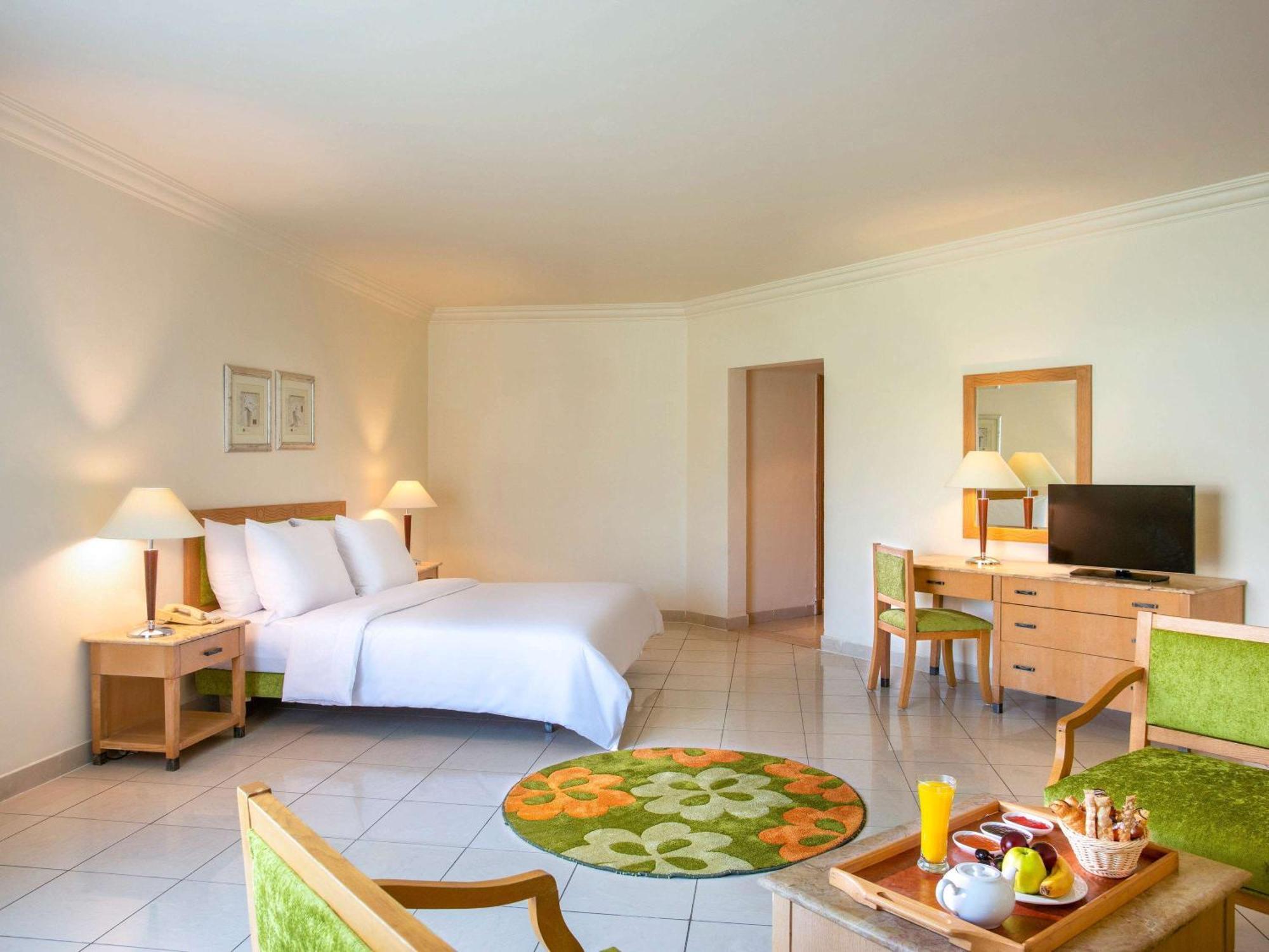 Movenpick Taba Resort & Spa Dış mekan fotoğraf The photo shows a spacious hotel room decorated in a light and airy style. There is a large bed with white linens against one wall. To the left, there are two lamps on nightstands. A small wooden desk and a flat-screen TV are positioned against anoth