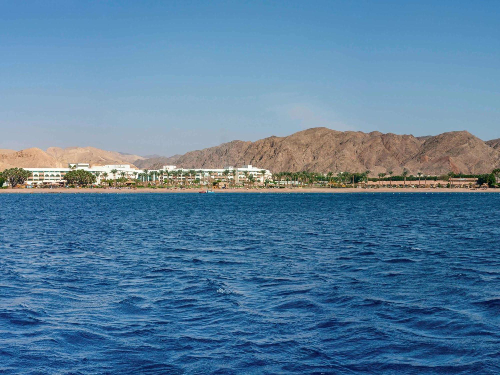 Movenpick Taba Resort & Spa Dış mekan fotoğraf The photo depicts a scenic view of a body of water in the foreground, with calm blue waves. In the background, there is a coastline featuring a resort or hotel lined with palm trees. Beyond the resort, rugged mountains rise, providing a beautiful nat