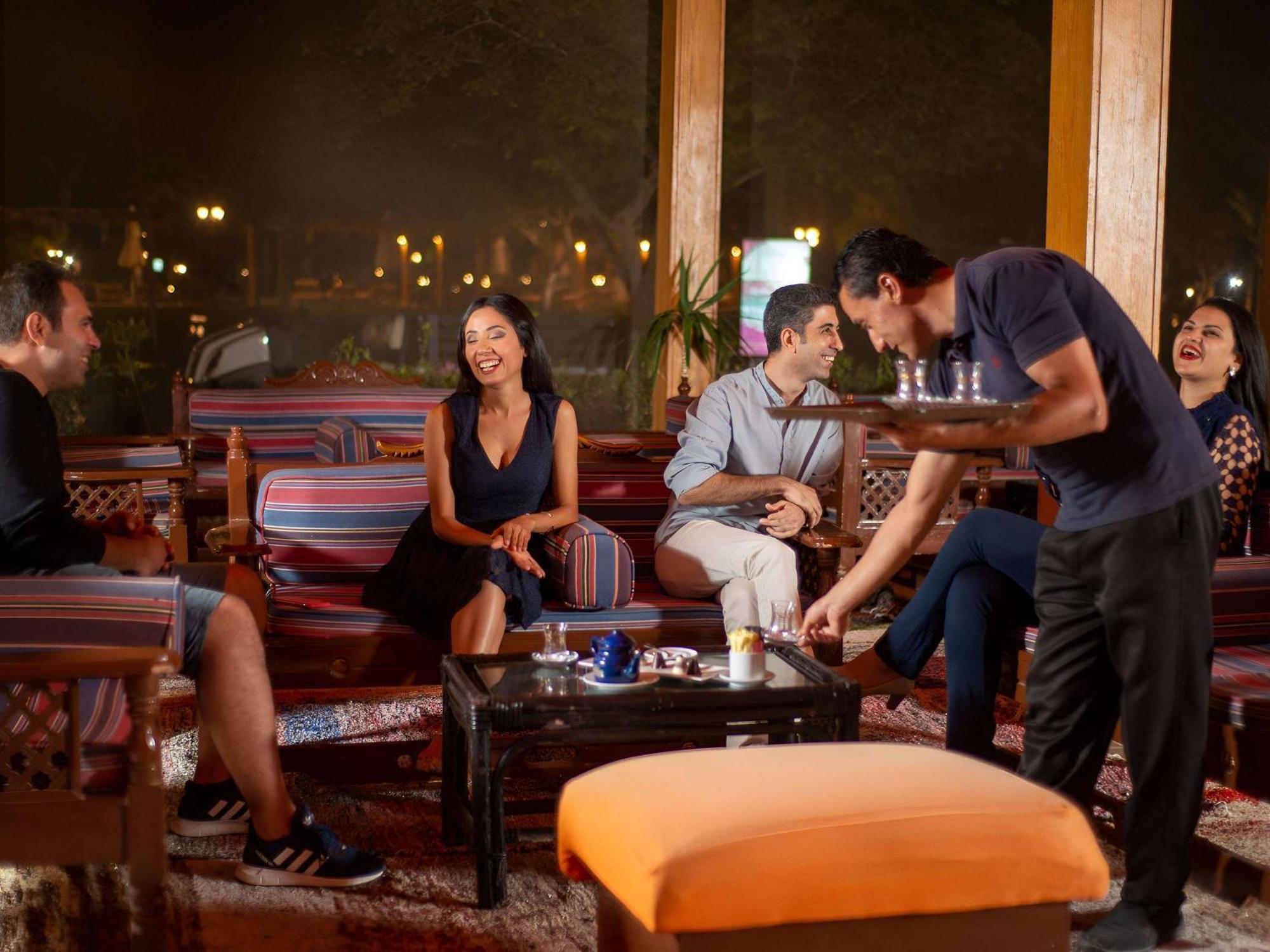 Movenpick Taba Resort & Spa Dış mekan fotoğraf The photo depicts a cozy indoor setting with a group of people enjoying each other's company. There are four individuals seated on comfortable furniture, engaged in conversation and laughter. A waiter is serving drinks from a tray to the group. The a