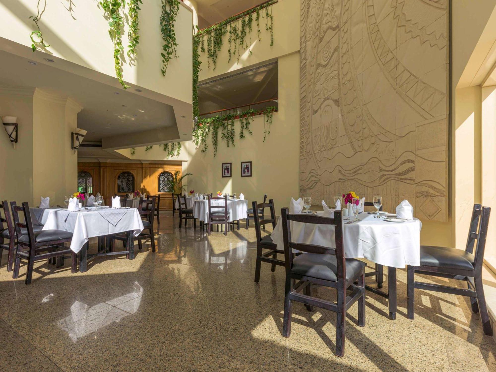 Movenpick Taba Resort & Spa Dış mekan fotoğraf The photo shows an indoor dining area of a restaurant. There are several tables, each covered with white tablecloths, and arranged with utensils and flower centerpieces. The space features a high ceiling, with plants hanging down from it, adding a to