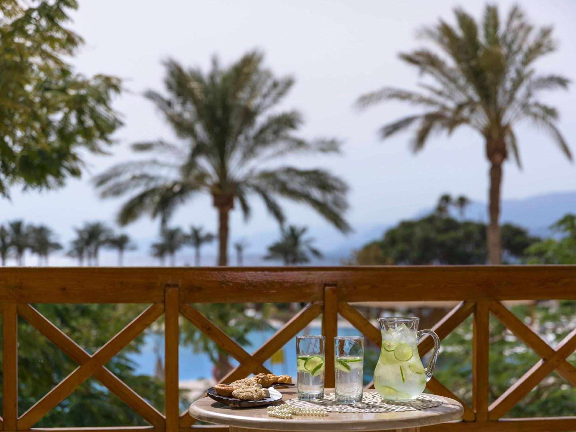 Movenpick Taba Resort & Spa Dış mekan fotoğraf The photo shows a scenic outdoor view with several tall palm trees in the background, indicating a tropical or warm environment. In the foreground, there's a small table with a tray that has two clear glasses filled with a light beverage, likely lemo