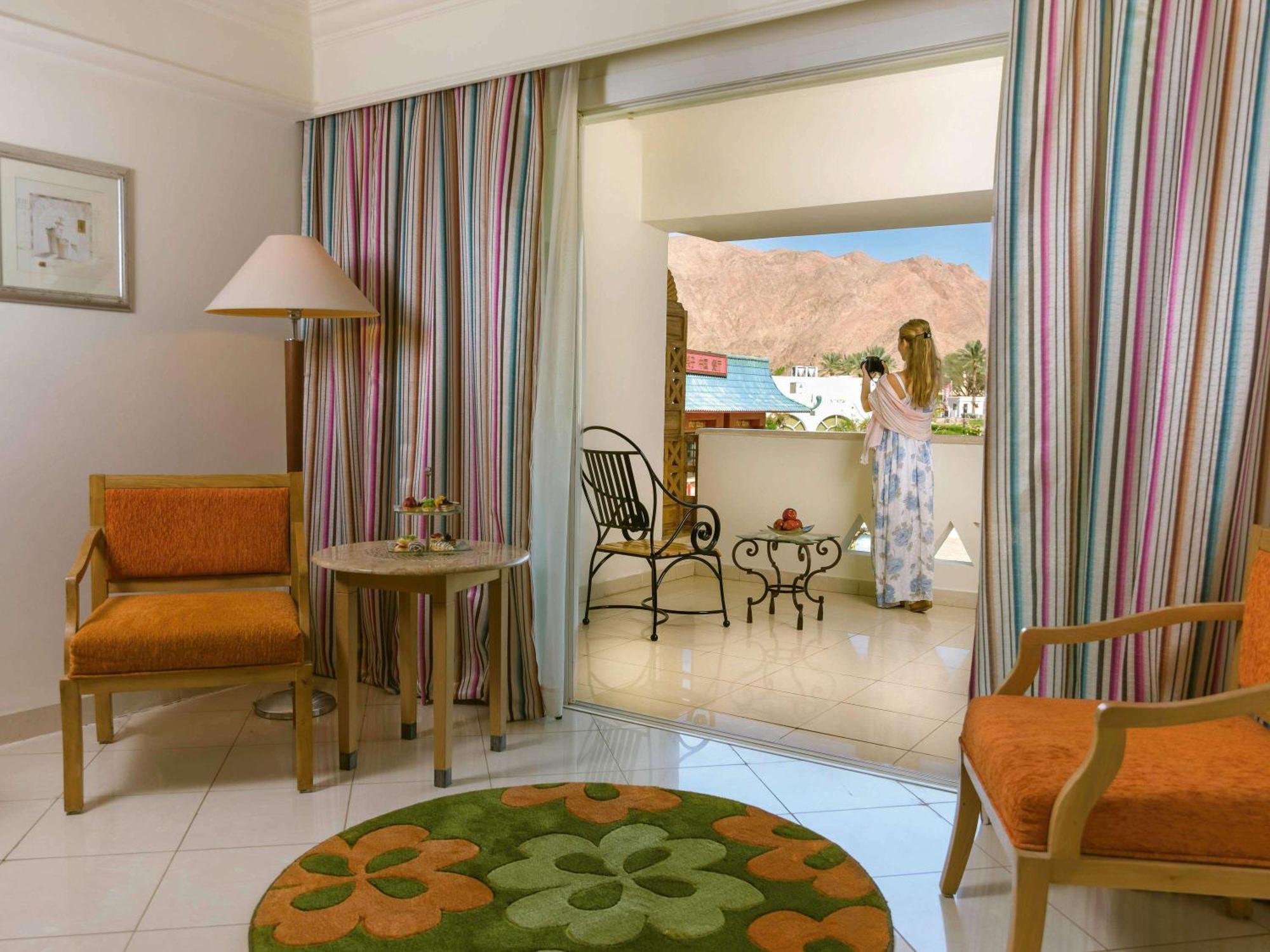 Movenpick Taba Resort & Spa Dış mekan fotoğraf The image shows a room with a cozy and inviting atmosphere. It features two orange chairs with wooden frames positioned around a small round table. There is a decorative rug on the floor with a floral pattern in green and orange. 

In the background,