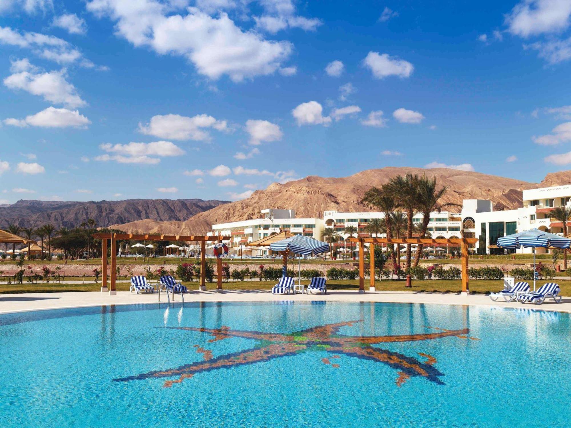 Movenpick Taba Resort & Spa Dış mekan fotoğraf The photo shows a tranquil swimming pool set against a backdrop of mountains. The pool's water is clear and reflects a beautiful blue sky with some fluffy clouds. Surrounding the pool are several lounge chairs, and the area appears to be part of a re