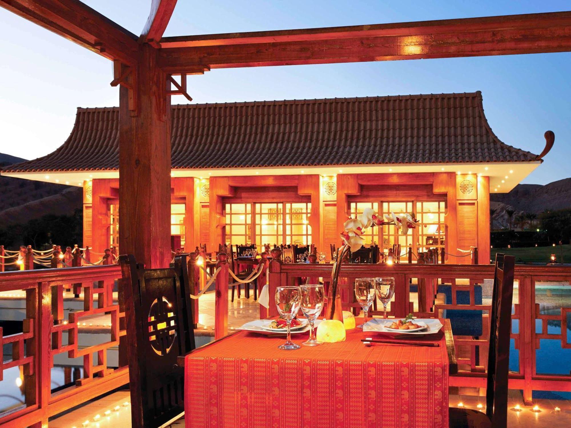 Movenpick Taba Resort & Spa Dış mekan fotoğraf The photo shows an outdoor dining setting at a beautifully designed restaurant with traditional architecture. The structure has a distinctive roof and is warmly illuminated, creating a cozy atmosphere at dusk. In the foreground, there is a table set 