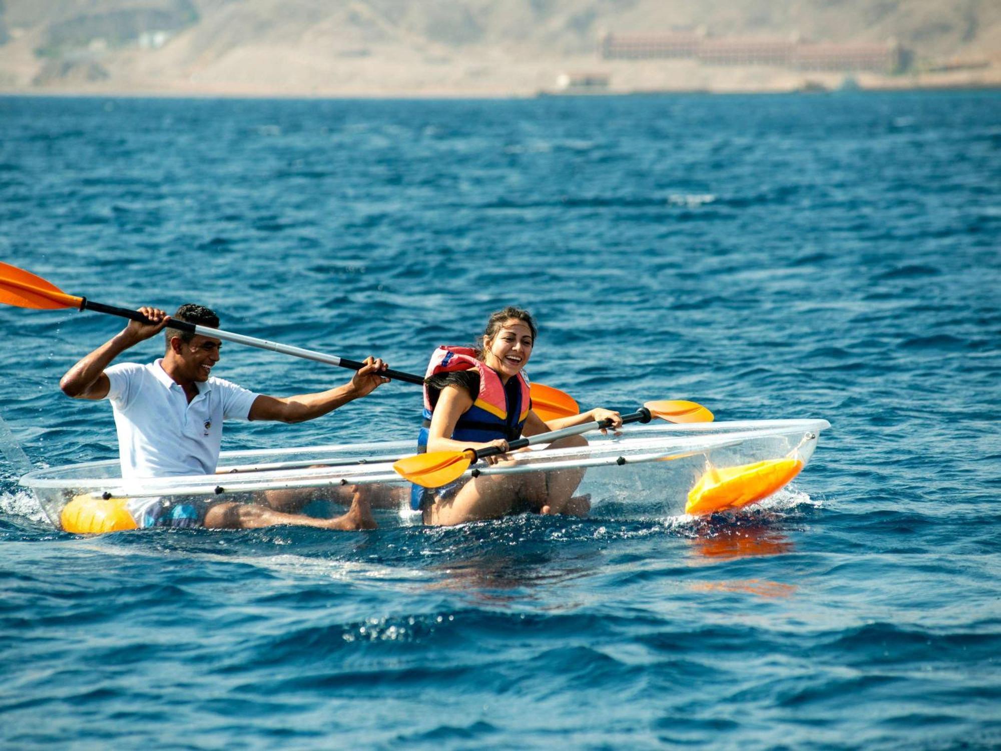 Movenpick Taba Resort & Spa Dış mekan fotoğraf The photo depicts two people paddling a kayak in a body of water. One person is seated at the front, laughing and enjoying the experience, while the other is at the back, actively using a paddle to steer. The water appears calm, and the surrounding e