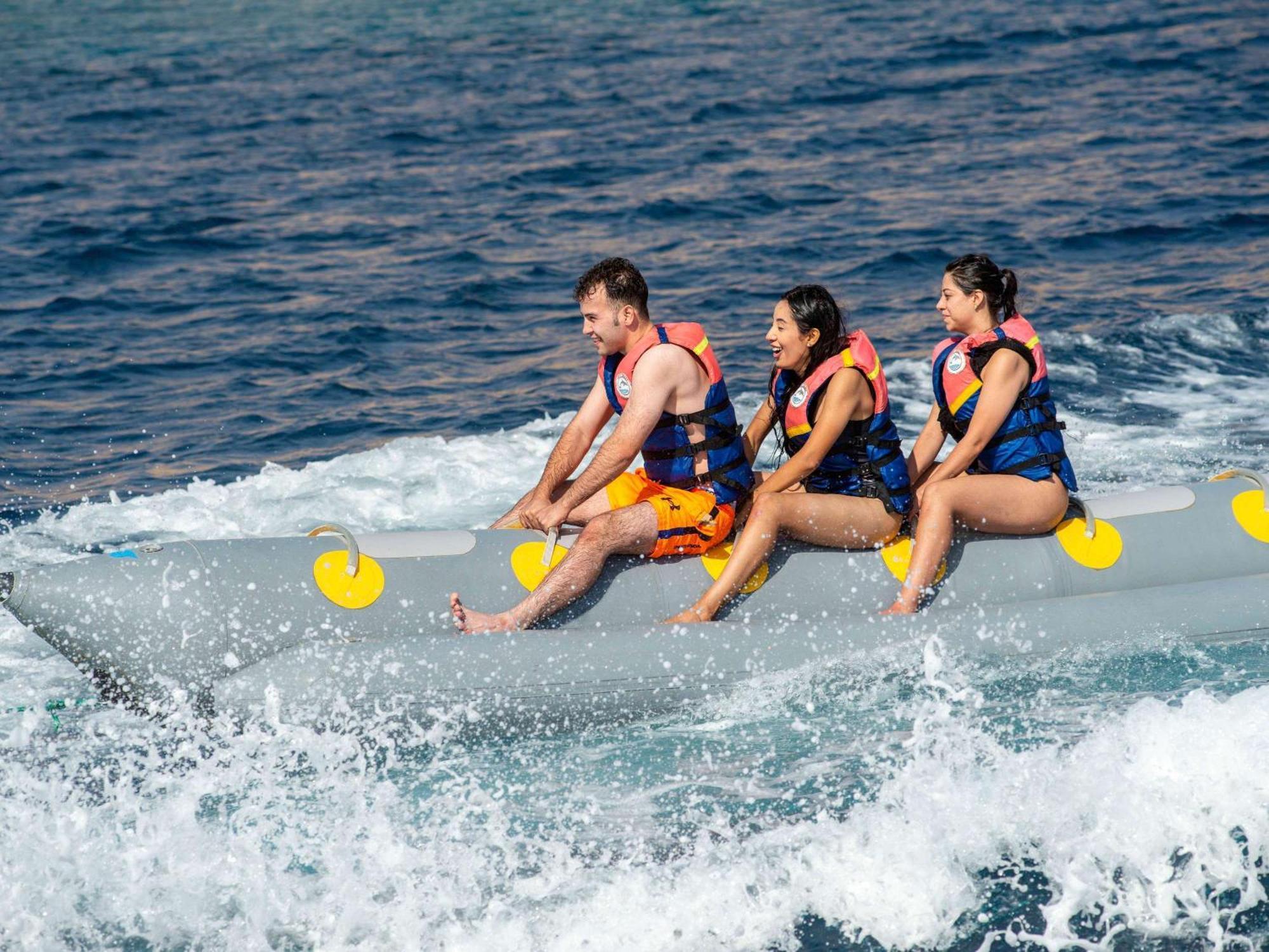 Movenpick Taba Resort & Spa Dış mekan fotoğraf The photo shows three people riding on a banana boat, which is a type of inflatable boat often used for recreational purposes on water. They are wearing life jackets and appear to be enjoying the ride as the boat speeds across the waves. The backgrou