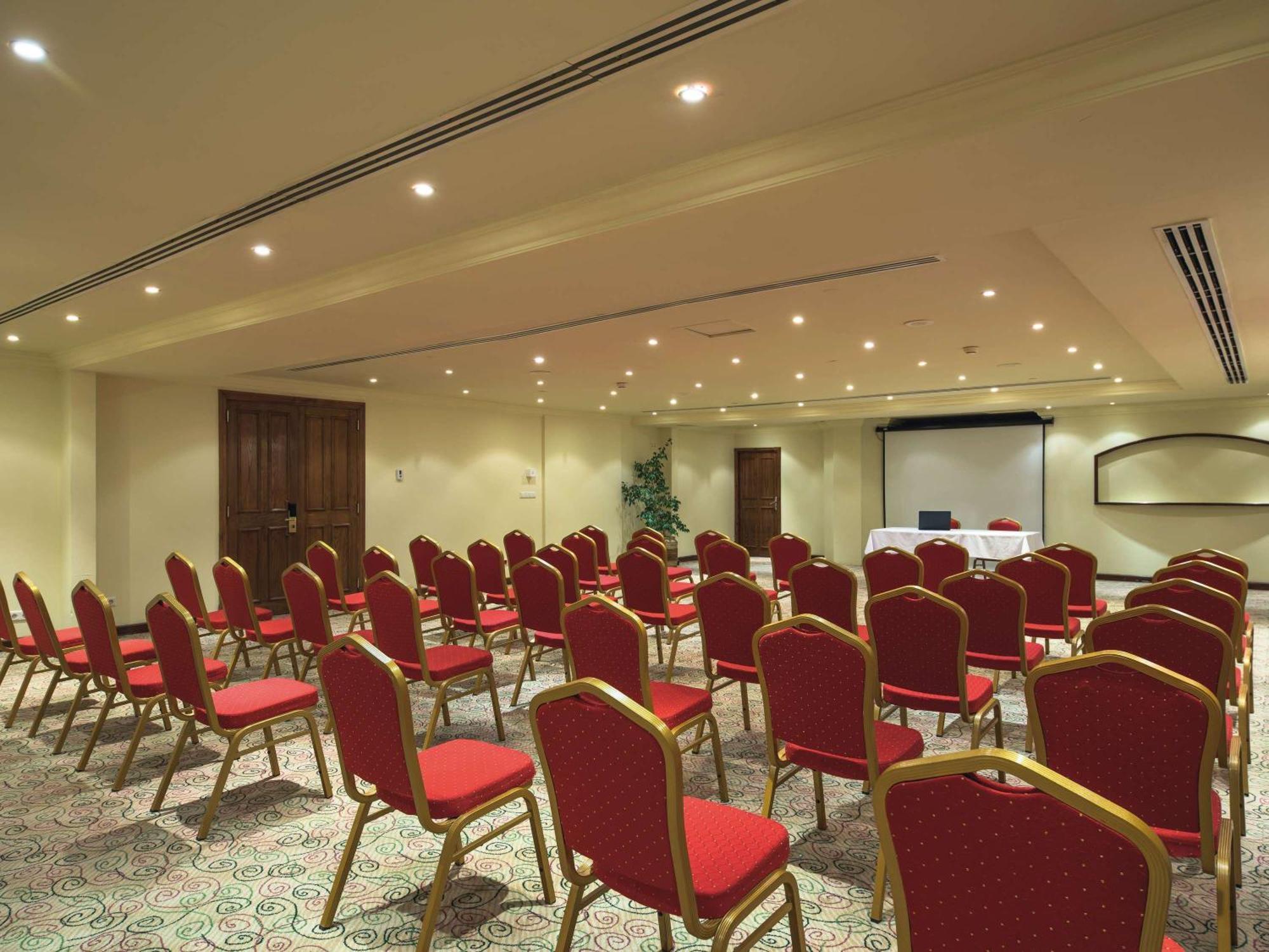 Movenpick Taba Resort & Spa Dış mekan fotoğraf The photo shows a conference room set up for a meeting or presentation. There are rows of red chairs arranged facing a front area where a presentation might take place. The room has neutral-colored walls and a carpet with a patterned design. Lighting