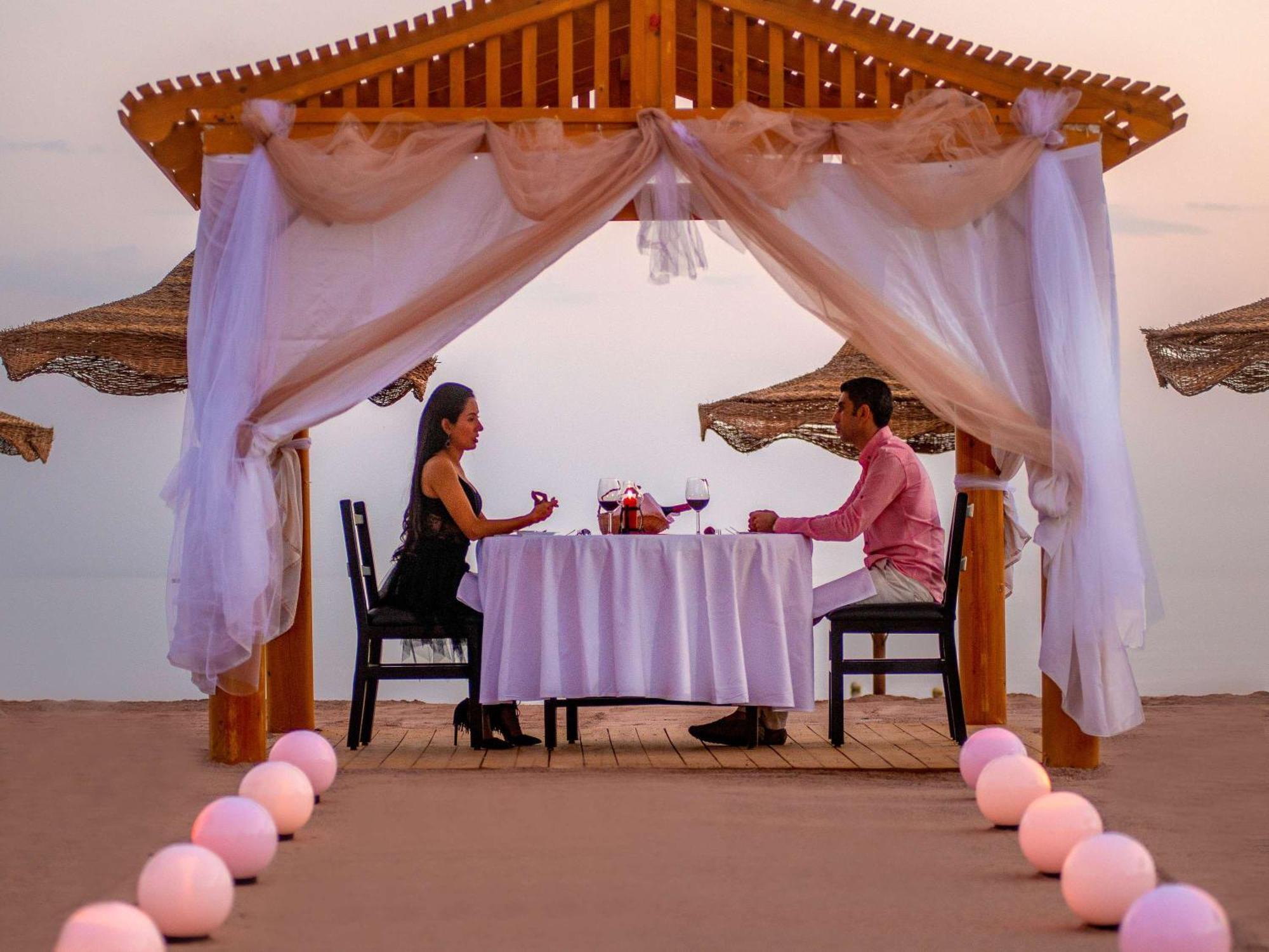 Movenpick Taba Resort & Spa Dış mekan fotoğraf The photo shows a romantic setting for two people dining together under a wooden pavilion. The pavilion is adorned with draped white fabric and soft decorations, creating an intimate atmosphere. A table is set with a white tablecloth, and there are t