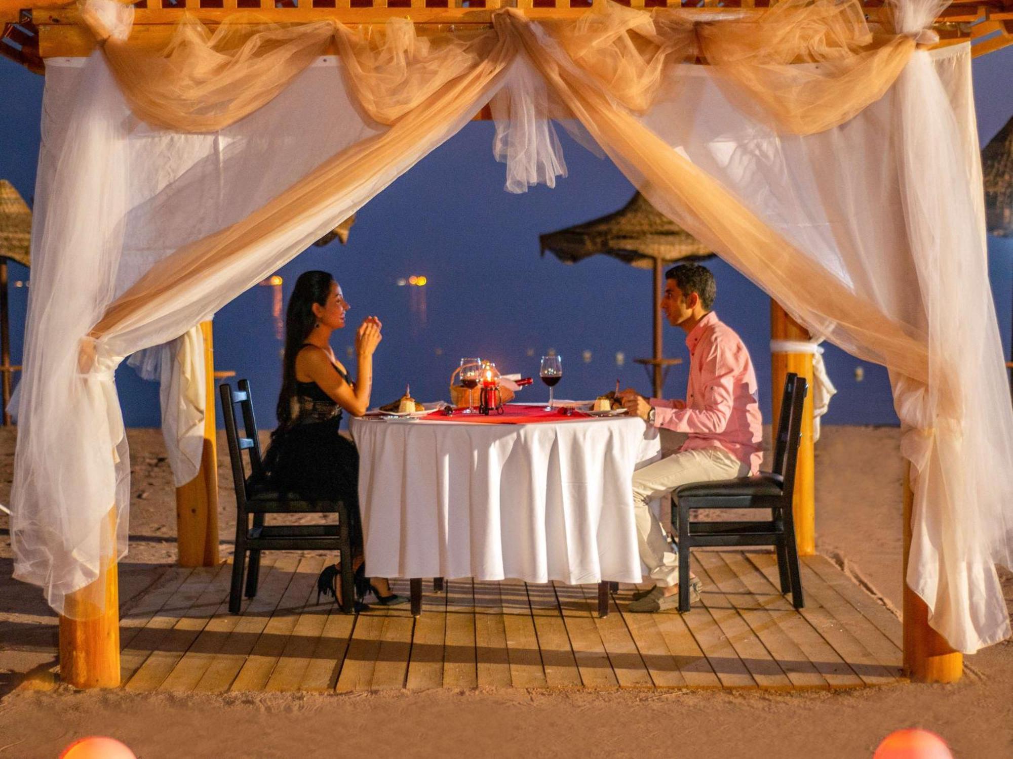 Movenpick Taba Resort & Spa Dış mekan fotoğraf The photo depicts a romantic dining setup on the beach. A table is elegantly set with a white tablecloth and decor, including wine glasses and a centerpiece. There are two people seated at the table: one in a black dress and the other in a pink shirt
