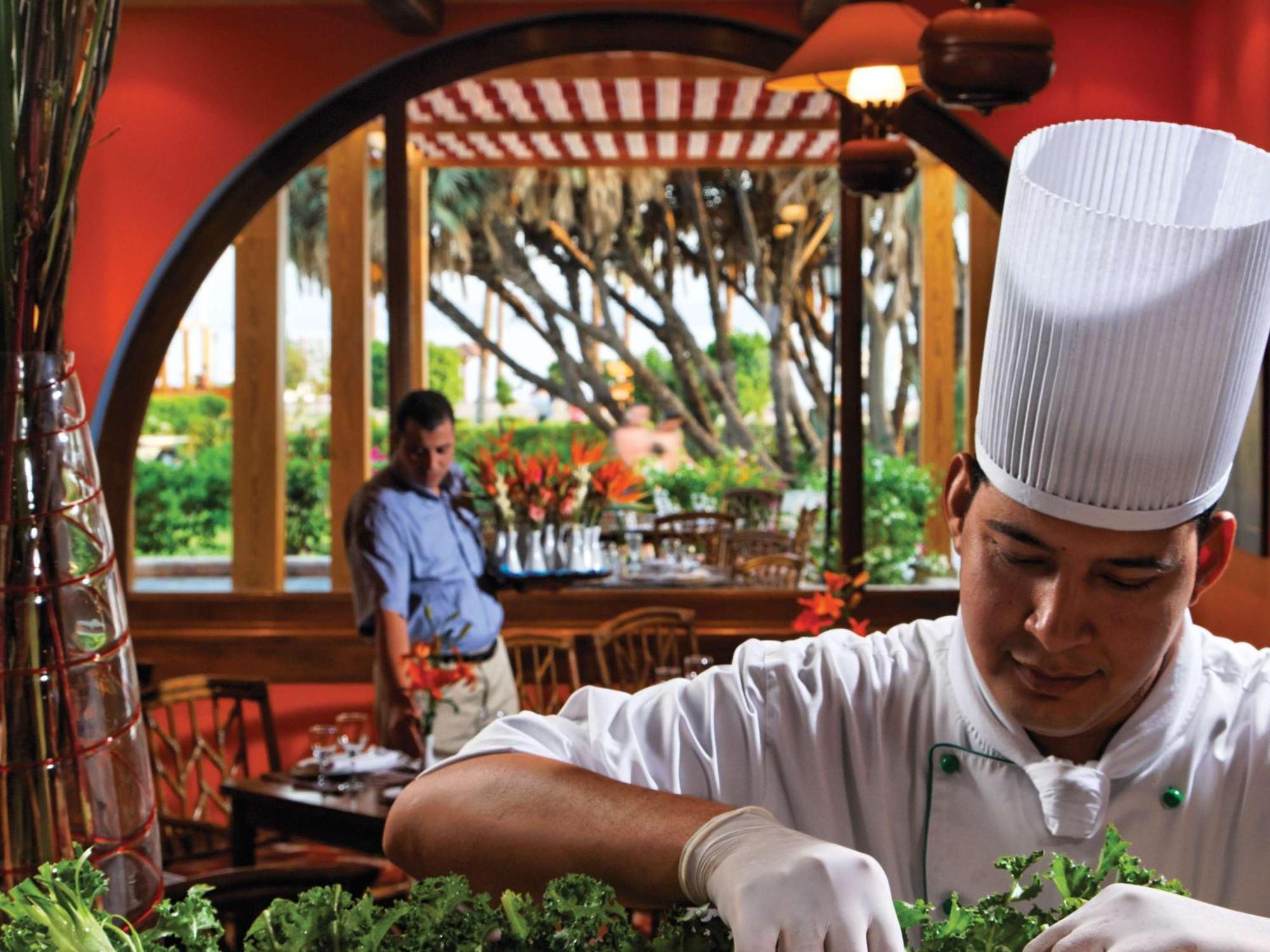 Movenpick Taba Resort & Spa Dış mekan fotoğraf The photo depicts a restaurant scene. In the foreground, a chef wearing a white hat and gloves is meticulously preparing ingredients, likely fresh vegetables or herbs, for a dish. His focus suggests a commitment to detail and quality in the culinary 