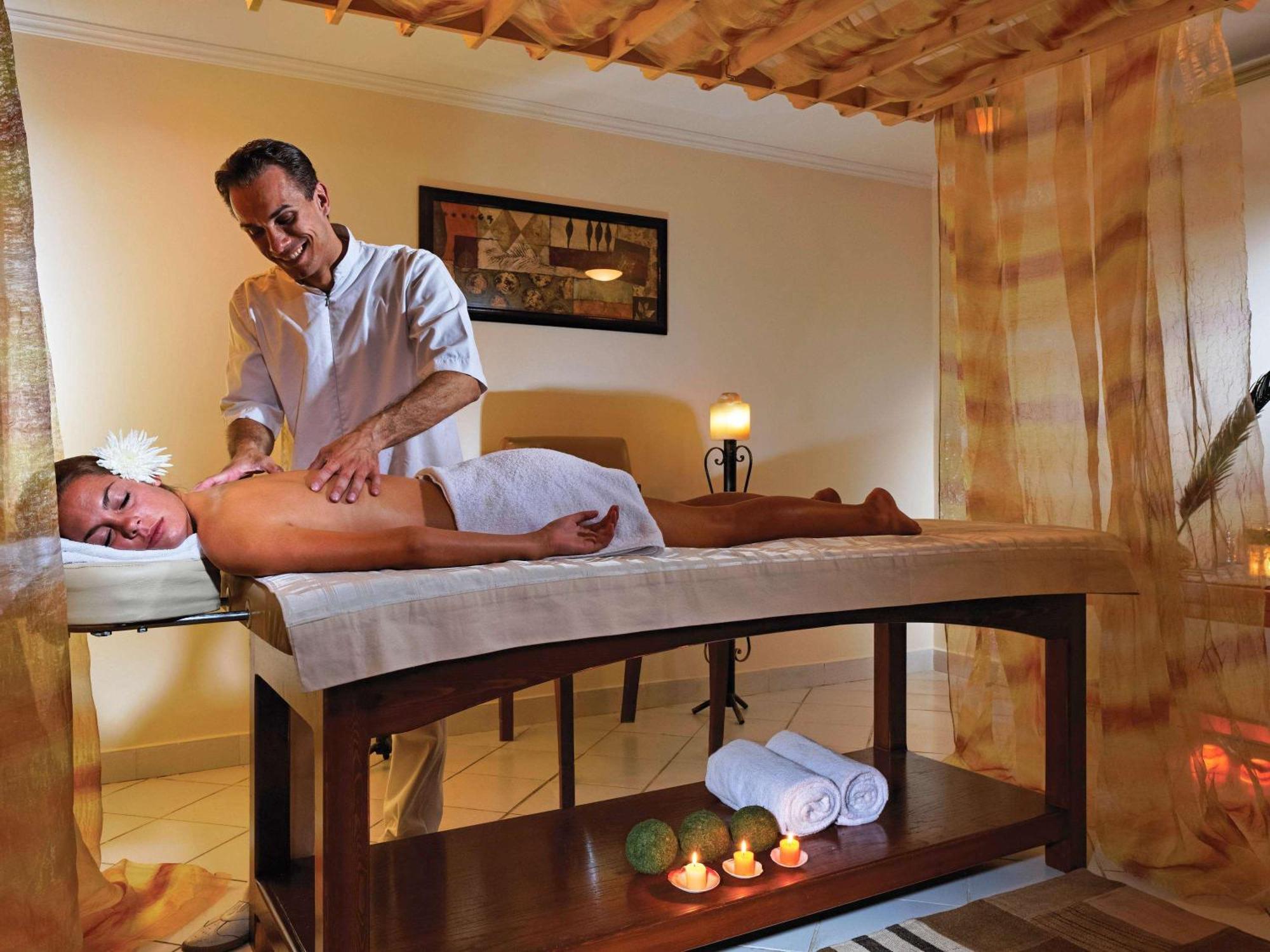 Movenpick Taba Resort & Spa Dış mekan fotoğraf The image depicts a serene spa setting where a therapist is giving a massage to a client lying on a massage table. The client is covered with a towel and has a flower in her hair, suggesting a relaxing and tranquil atmosphere. The room is softly lit 