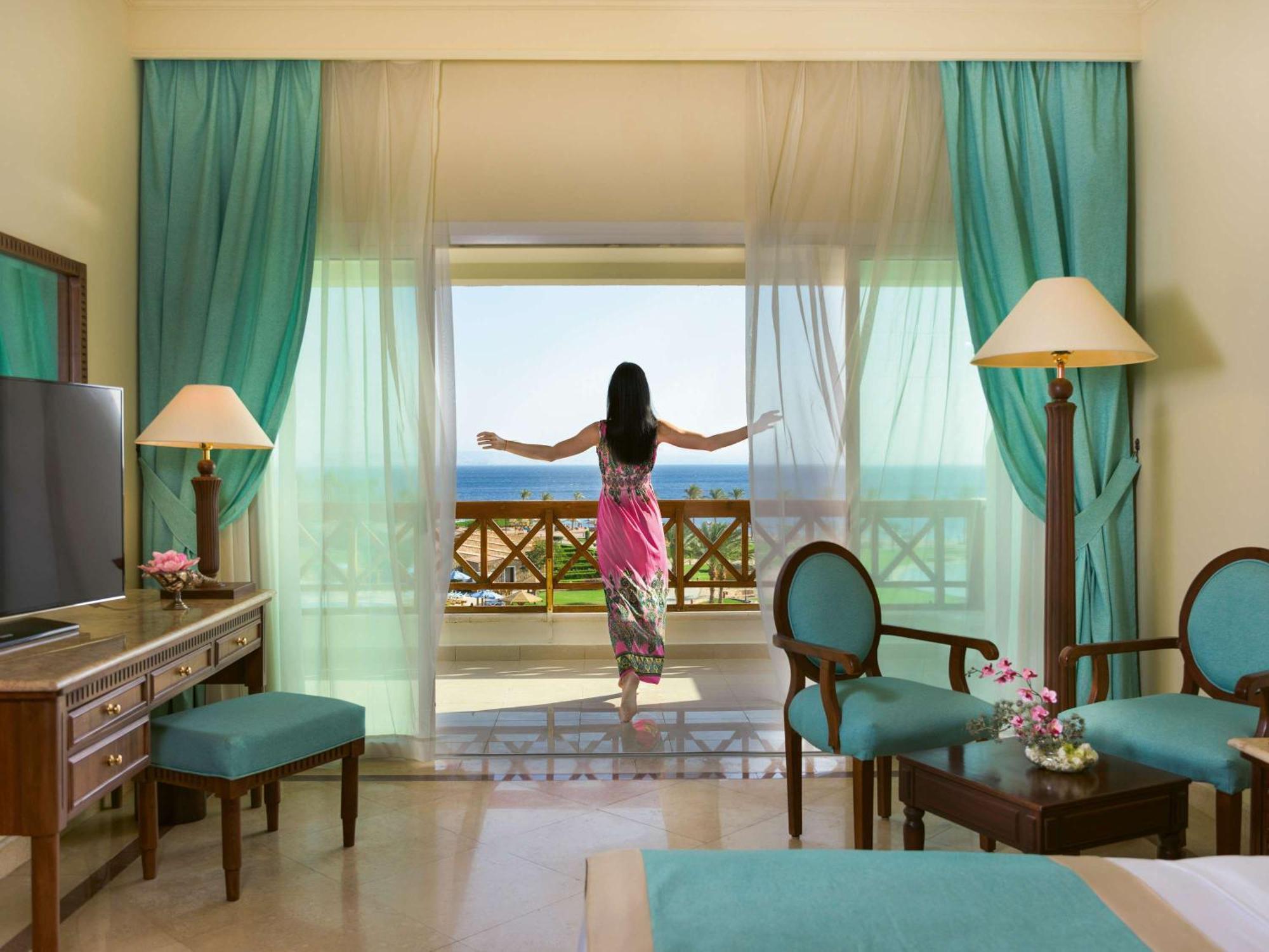 Movenpick Taba Resort & Spa Dış mekan fotoğraf The photo shows a serene hotel room with a person standing by a large glass door, enjoying a beautiful view. The room features light-colored walls, turquoise curtains, and elegant furniture, including a TV, a wooden desk, and chairs with matching uph