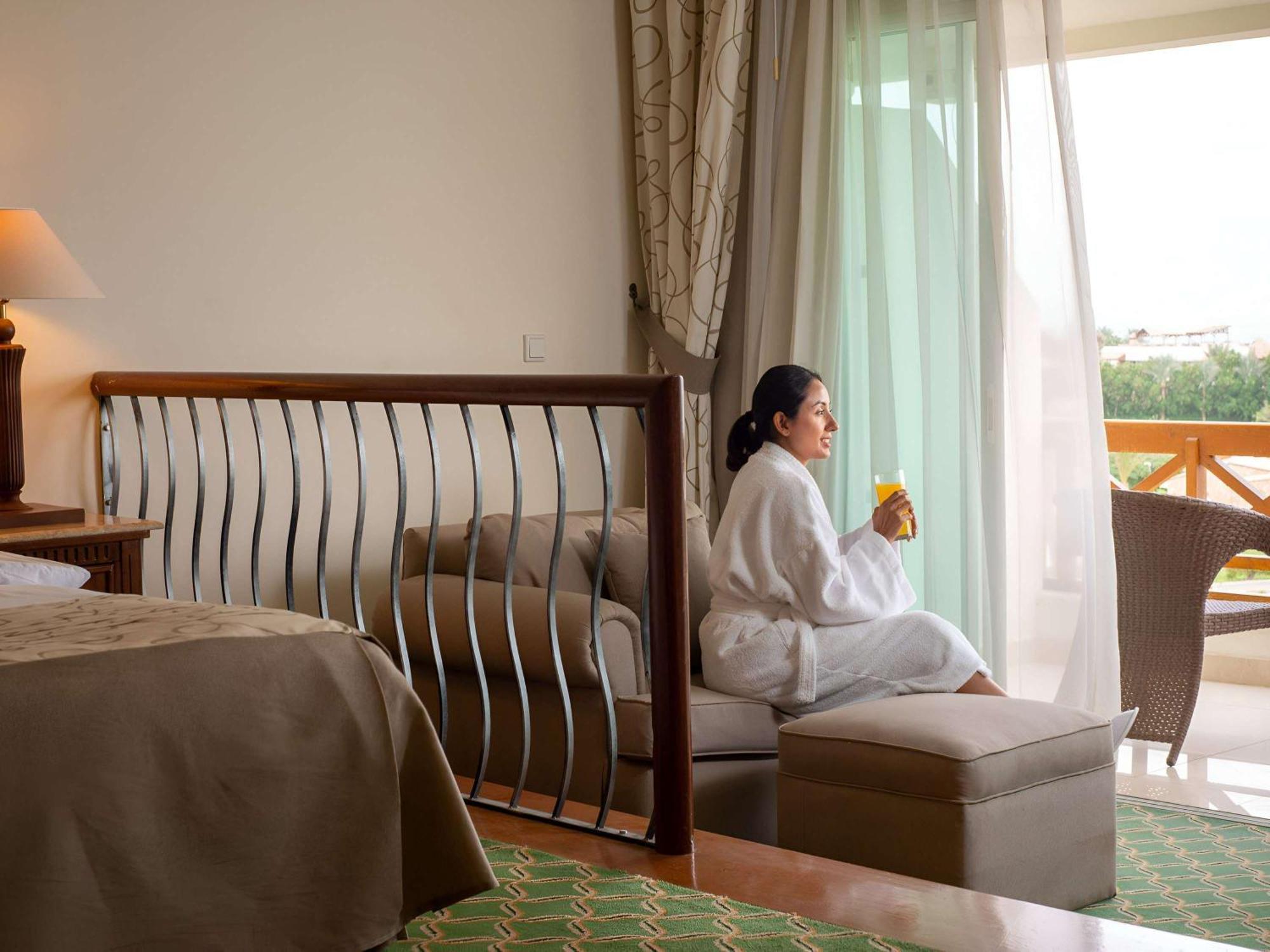 Movenpick Taba Resort & Spa Dış mekan fotoğraf The photo shows a well-decorated hotel room with a cozy atmosphere. In the foreground, there is a bed with neutral-toned bedding and a decorative headboard. A woman is seated on a cushioned ottoman, wearing a white bathrobe and holding a drink. She i