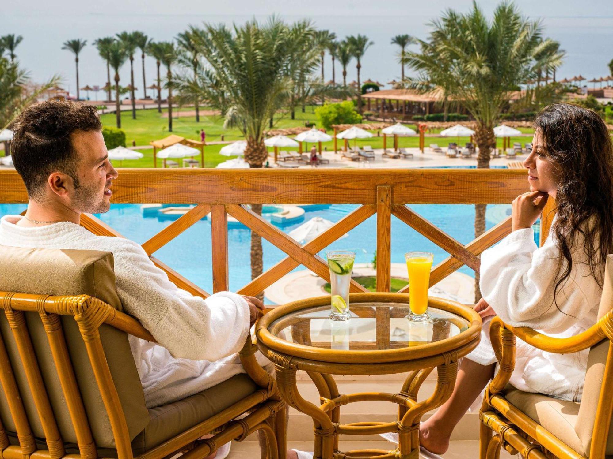 Movenpick Taba Resort & Spa Dış mekan fotoğraf The photo depicts a couple sitting on a balcony overlooking a scenic view. They are relaxing in bathrobes and seated at a small table. Each person has a drink; one has a yellow beverage and the other has a green one. In the background, there are palm