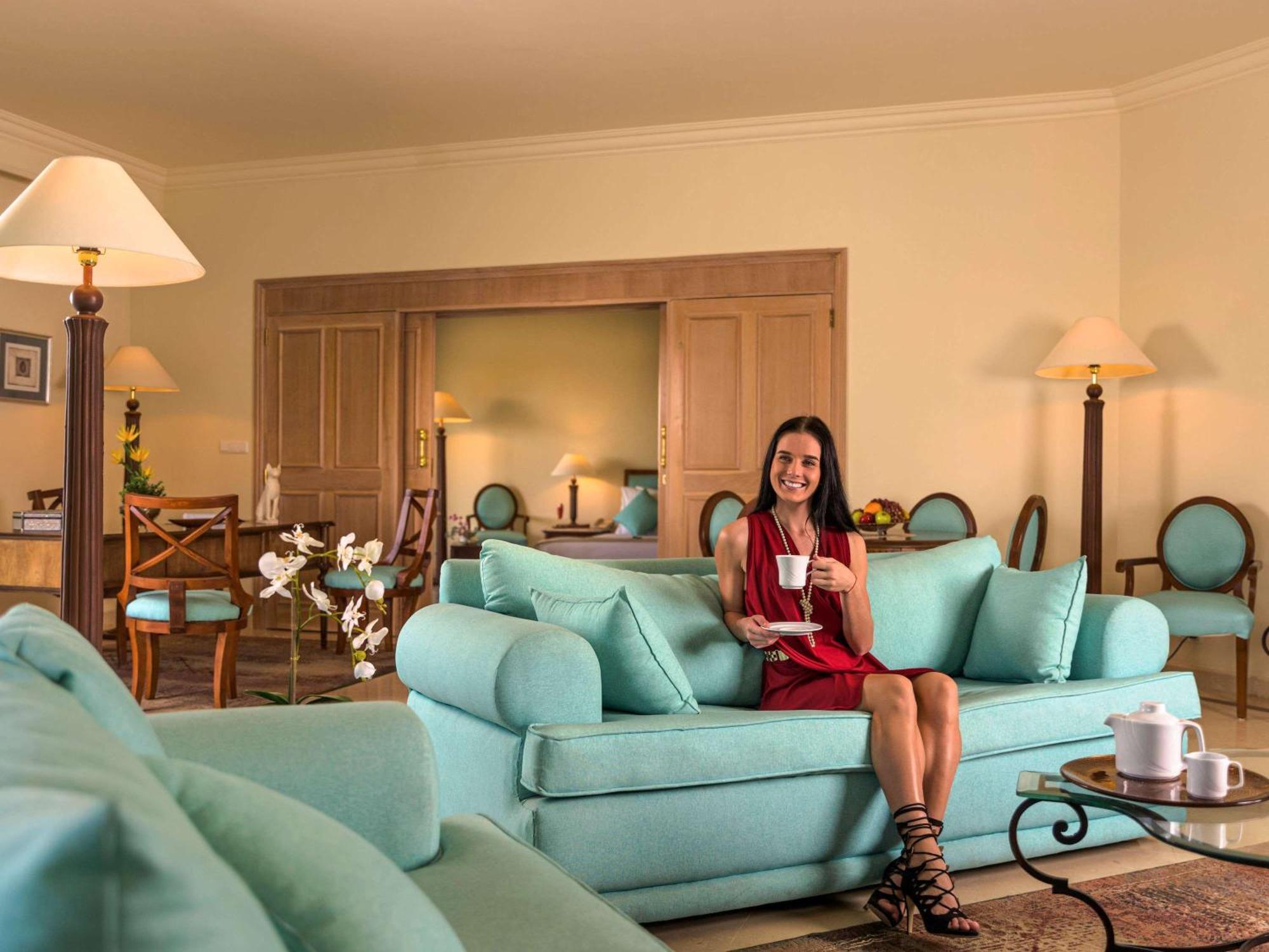 Movenpick Taba Resort & Spa Dış mekan fotoğraf The photo depicts a cozy and elegantly decorated living space. A woman with a cheerful expression is seated on a light teal sofa, holding a cup of tea or coffee. She is dressed in a red dress and appears relaxed. Surrounding her are tastefully arrang