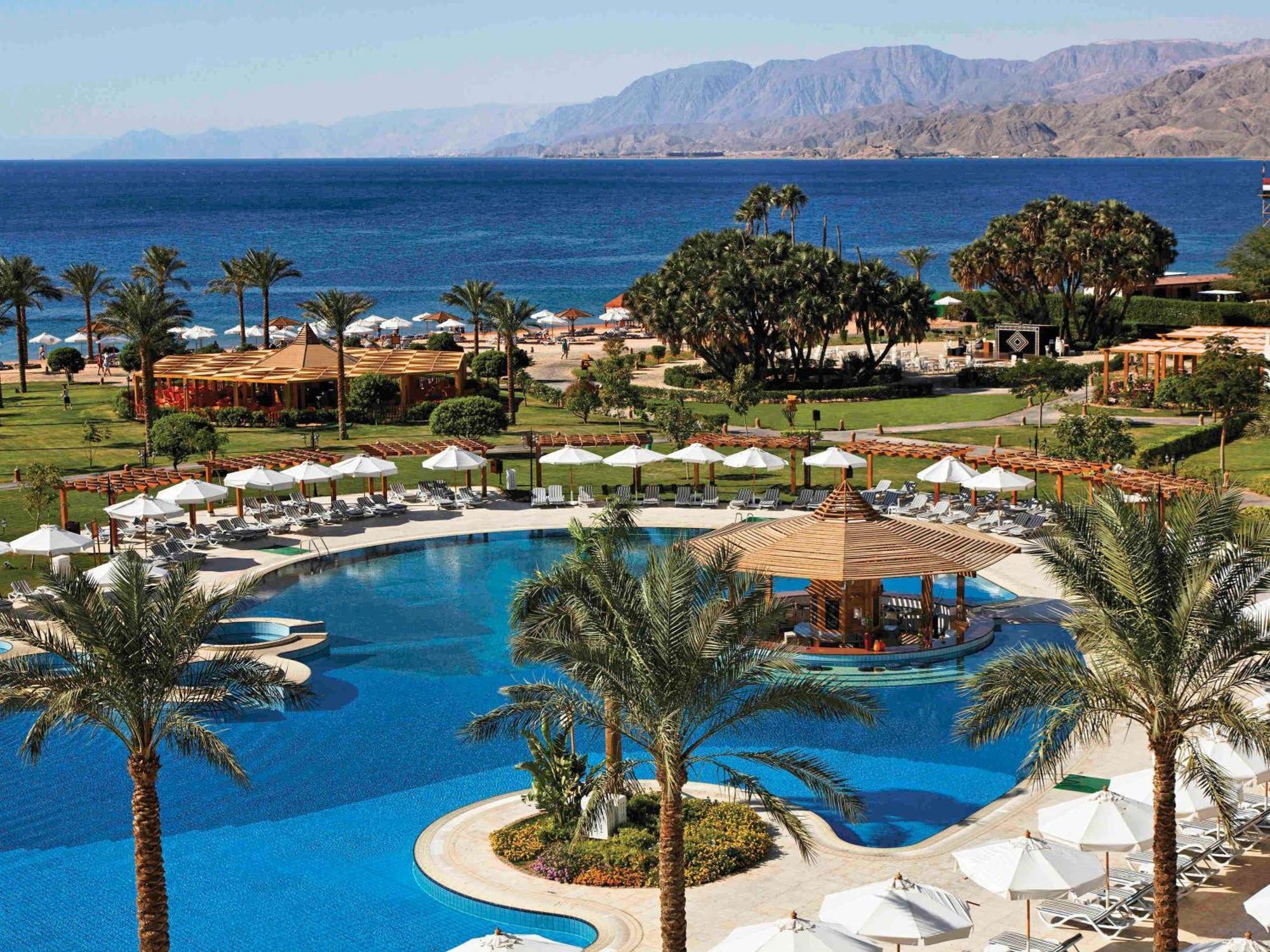 Movenpick Taba Resort & Spa Dış mekan fotoğraf The photo shows a luxurious resort overlooking a large, blue swimming pool. Surrounding the pool are palm trees and lounge chairs with white umbrellas, creating a relaxing atmosphere. In the background, the ocean can be seen, with a mountainous lands